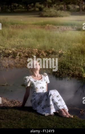 Une femme blonde dans une robe florale est assis à un étang, à profiter du soleil Banque D'Images