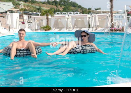 Young couple holding hands dans une piscine qu'elles flottent côte à côte sur les tubes en caoutchouc le soleil brille sur les vacances d'été. Banque D'Images