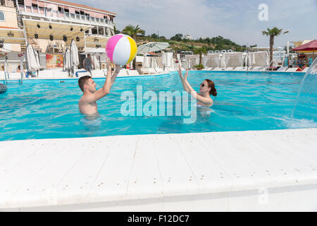 Jeune couple de lancer un ballon de plage à l'autre comme ils s'ébattre autour dans un endroit invitant bleu piscine d''un summer resort Banque D'Images