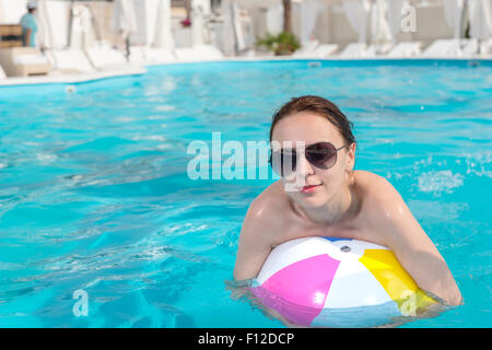 Close up jolie jeune femme avec des lunettes, serrant un ballon de plage dans la piscine et souriant à la caméra. Banque D'Images