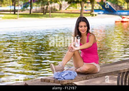 Brunette model wearing pink top et short blanc se détendre dans l'environnement du parc, assis sur un banc à côté du lac en utilisant un insectifuge spray Banque D'Images