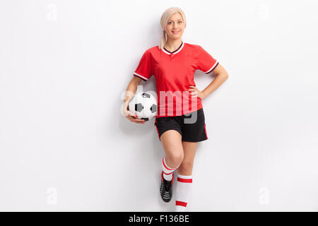 Le joueur de soccer féminin dans un maillot rouge et un short noir tenant une balle et appuyé contre un mur Banque D'Images