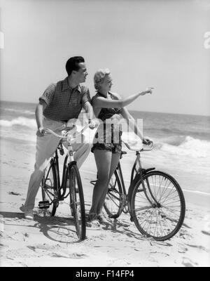 1930 HOMME FEMME COUPLE WALKING ON BEACH FEMME pointant à l'HORIZON Banque D'Images