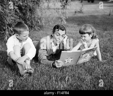 1950 HOMME PÈRE ALLONGÉ DANS L'HERBE LECTURE LIVRE À SON FILS ET SA FILLE Banque D'Images