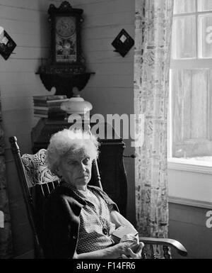 1960 SAD LONELY WOMAN SITTING IN ROCKING CHAIR ILLUMINÉE PAR LA LUMIÈRE DE LA FENÊTRE Banque D'Images