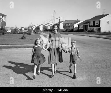 1950 SMILING HAPPY MOTHER HOLDING HANDS FILS ET FILLE DE MARCHER À TRAVERS LE DÉVELOPPEMENT DU LOGEMENT DE BANLIEUE STREET LOOKING AT CAMERA Banque D'Images