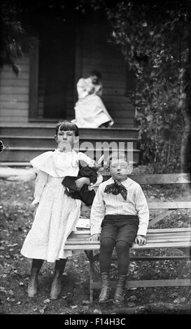 Années 1900 PORTRAIT JEUNE FILLE GARÇON ASSIS SUR UN BANC EN FACE DE LA MAISON À LA CAMÉRA À WOMAN IN BACKGROUND ON PORCH Banque D'Images