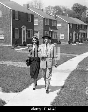 1950 HOMME FEMME COUPLE bras dessus bras dessous MARCHANT DANS LA BANLIEUE DE TROTTOIR RUE DE MAISONS EN Briques de construction récente Banque D'Images