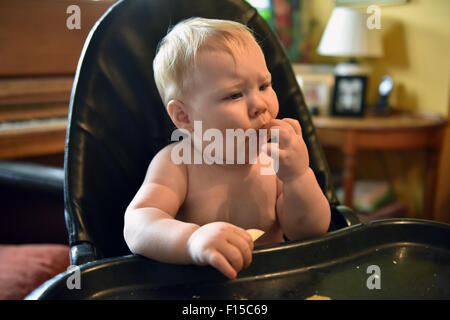 Un bébé 11 mois assis sur une chaise haute de manger une tranche de pomme. Banque D'Images