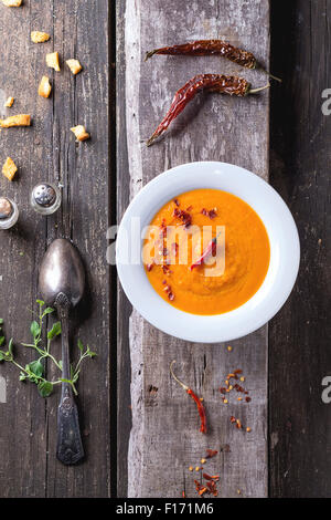 Plaque blanche de soupe crème de carottes avec red hot chili pepper. Plus de vieille table en bois avec vintage cuillère, poivrons, sel et croûtons Banque D'Images