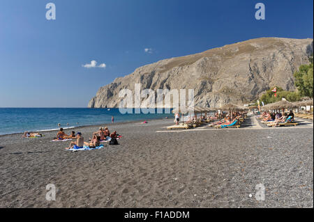 Les touristes à prendre le soleil sur la plage de sable noir de Kamari, Santorini, Grèce. Banque D'Images