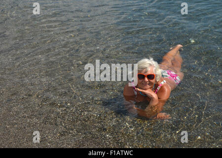 Portrait de senior lady c'est couché dans l'eau peu profonde de la mer de galets de plage. Banque D'Images