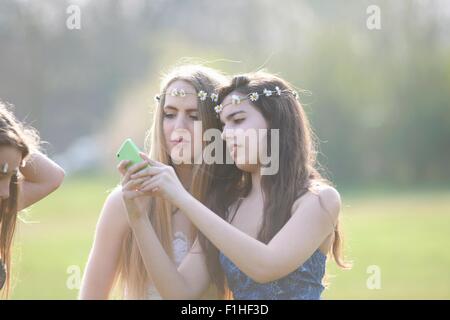 Deux jeunes filles portant des coiffes en chaîne smartphone lecture textes dans park Banque D'Images