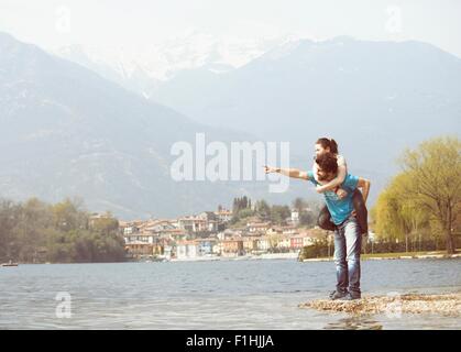 Young man giving girlfriend piggyback sur lakeside, lac Mergozzo, Verbania, Piemonte, Italie Banque D'Images