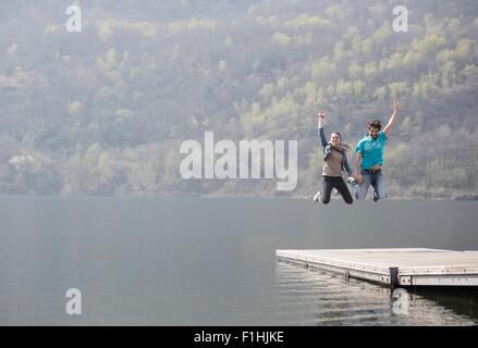 Jeune couple jumping mid air sur la jetée au lac Mergozzo, Verbania, Piemonte, Italie Banque D'Images