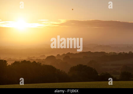 Un ballon à air chaud passe sur les arbres et les collines couvertes de brume au coucher du soleil, l'est du Devon, Angleterre. Banque D'Images