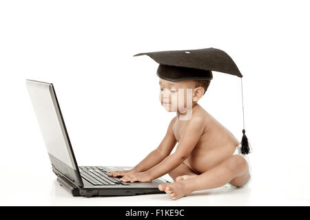 Un bébé assis devant un fond blanc, portant une graduation cap et de jouer sur un ordinateur portable. Banque D'Images