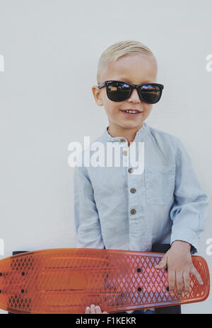 Petit garçon dans le quartier branché de grandes lunettes emprunté à un parent l'article saisissant un skateboard regardant la caméra avec un c Banque D'Images