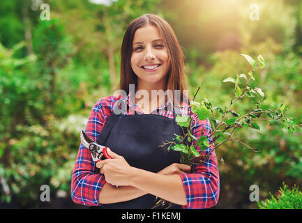 Souriante jeune femme élagage jardinier les plantes debout dans un jardin verdoyant avec une évaluation annuelle des sécateurs dans sa main smiling at Banque D'Images