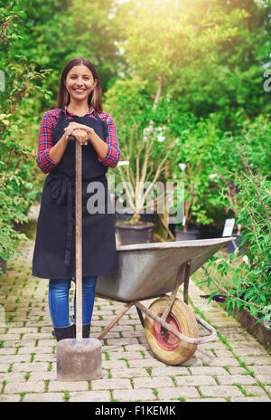 Jeune femme de pépinière repiquage debout avec une plante en pot dans une brouette dans la serre s'appuyant sur un spa Banque D'Images