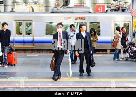 La gare d'Osaka City. Deux hommes-salaire japonais en costumes en attente de trajet du matin sur la plate-forme, en face. Les deux exerçant son porte-documents, l'un avec un masque. Banque D'Images