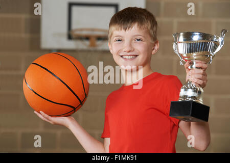 Boy Holding Basket-ball et le trophée en gymnase de l'école Banque D'Images