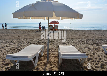 Chaises longues et parasol sur la plage de Playa San Juan, Alicante, avec des gens qui marchent sur le rivage en début de matinée. Banque D'Images