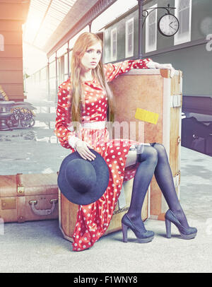 La fille assise sur la valise en attente à la gare ferroviaire de retro Banque D'Images