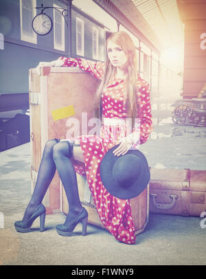 La fille assise sur la valise en attente à la gare ferroviaire de retro Banque D'Images