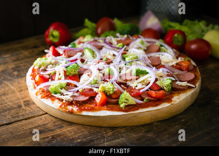 Pizza à croûte mince avec saucisses, tomates, poivrons, brocoli et fromage sur planche de bois rond Banque D'Images