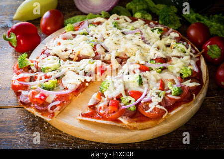 Pizza à croûte mince avec saucisses, tomates, poivrons, brocoli et fromage sur planche de bois rond Banque D'Images