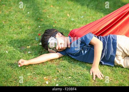 Garçon portant ces lunettes et cape gisant sur le côté de l'herbe, position de vol Banque D'Images