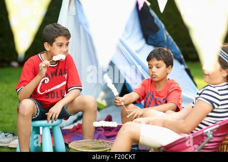 Girl et frères de manger les guimauves grillées devant des tente dans jardin Banque D'Images