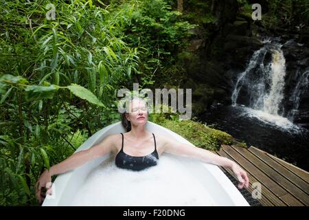 Mature Woman relaxing in bubble bath en face de cascade à eco retreat Banque D'Images