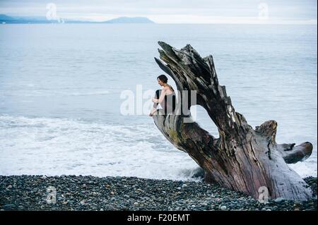 Mature Woman sitting on grand tronc de bois flotté at beach Banque D'Images