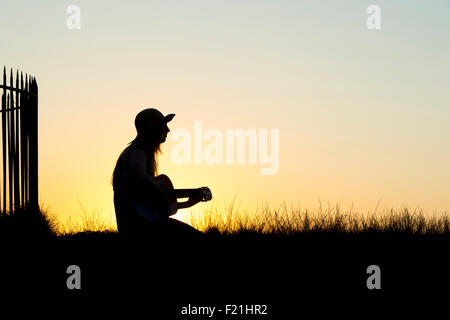 Adolescente assis jouant de la guitare au coucher du soleil. Silhouette Banque D'Images