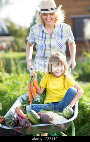 Mère et enfant dans un jardin intérieur. Enfant assis dans la brouette avec des légumes de la récolte. Les légumes biologiques sains pour les enfants. Banque D'Images