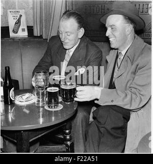 Photo historique datant des années 1950, deux hommes adapté, l'un avec un imperméable et portant un chapeau, assis ensemble dans un pub et profiter de ses pintes et bouteilles de bière blonde Wilsons, Manchester, Royaume-Uni. Banque D'Images