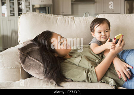 Femme allongée sur un canapé, smiling, câlins avec son jeune fils et à la recherche à un téléphone cellulaire. Banque D'Images