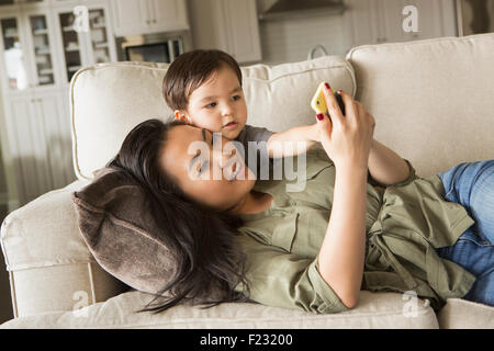 Femme allongée sur un canapé, smiling, câlins avec son jeune fils et à la recherche à un téléphone cellulaire. Banque D'Images