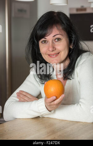 Happy woman holding une orange, à la recherche dans l'appareil photo dans la cuisine Banque D'Images