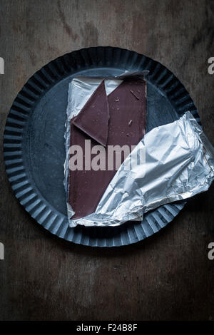 À mi-chemin unwrapped barre de chocolat avec une pièce cassée sur la plaque d'étain rustique sur fond de bois. Vue d'en haut Banque D'Images