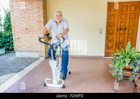 Personnes âgées de sexe masculin ne cesse de monter par l'octogénaire doing exercise bike sur le patio de la chambre Banque D'Images