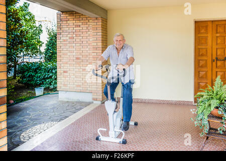 Personnes âgées de sexe masculin ne cesse de monter par l'octogénaire doing exercise bike sur le patio de la chambre Banque D'Images