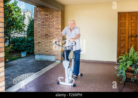 Personnes âgées de sexe masculin ne cesse de monter par l'octogénaire doing exercise bike sur le patio de la chambre Banque D'Images