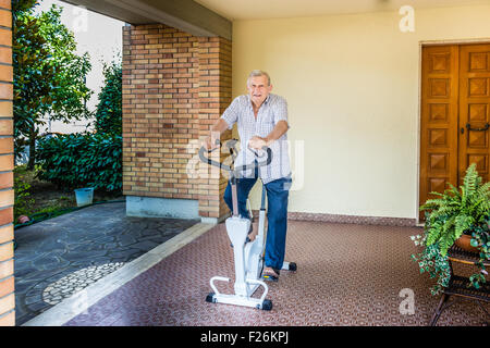 Personnes âgées de sexe masculin ne cesse de monter par l'octogénaire doing exercise bike sur le patio de la chambre Banque D'Images