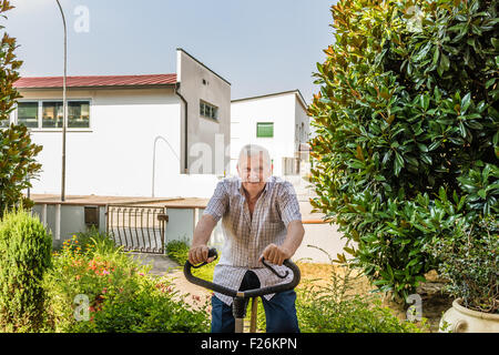 Personnes âgées de sexe masculin ne cesse de monter par l'octogénaire doing exercise bike sur le jardin de la maison Banque D'Images