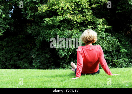 Vue arrière de femme d'âge moyen portant couleur rose corail top et pantalon bleu sitting on grass outdoors Banque D'Images