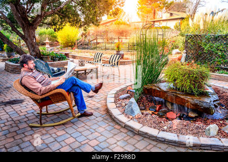 L'homme est en train de lire un journal sur un patio dans un jardin agréable Banque D'Images