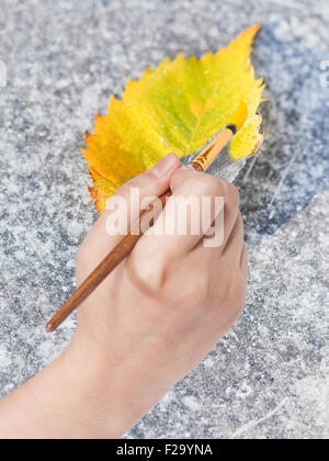 Concept nature - la main avec des peintures au pinceau dans la couleur jaune des feuilles tombées sur le trottoir gelé en automne Banque D'Images